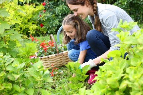 A woman with long brown hair, dressed in a light grey top and dark blue jeans, is leaning over to assist a young girl with long brown hair, wearing a bright blue shirt, as they work together in a lush garden. The girl is kneeling on the ground, holding a small watering can, while the woman guides her in tending to the vibrant plants. They are surrounded by various green foliage, including leafy shrubs and flowering plants with red blooms, which occupy the foreground and background. The garden features a well-maintained grass lawn with a slightly uneven texture, bordered by a mixture of flowering plants and dense greenery. Sunlight filters through the foliage, casting soft light on the scene, indicating a clear, pleasant day. This outdoor setting appears to be part of a residential backyard designed for family gardening activities. The scene highlights gardening, planting, and outdoor care, reflecting services offered by Gardening Services Southfields in the Southfields area, SW19 postcode, nearby London.