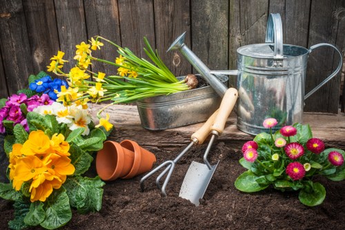 The image shows a garden scene with freshly turned dark soil in the foreground, featuring various gardening tools and decorative potted plants. To the left, there are vibrant, colorful flowers including yellow primroses, purple pansies, and white blooms, arranged among lush green foliage. In the center, a small metal garden trowel with a beige handle, a hand rake, and a hand fork are positioned on the soil, suggesting recent planting activity. Behind these tools, a metallic watering can and a rectangular metal container hold a bunch of yellow daffodils with long green stems, adding a touch of springtime freshness. To the right, a cluster of pink and yellow daisies in a pink flower pot provides a lively contrast against the dark soil background. The scene is set against a weathered wooden fence, indicating an outdoor garden workspace, with natural light illuminating the vibrant colors and textures of the plants and gardening equipment, which collectively support gardening services in Southfields.